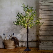 Dekorativer künstlicher Olivenbaum mit warmweißer LED-Lichterkette und schwarzem Standfuß, frontal in Innenraumkulisse vor rustikaler Wand und Holzboden.