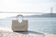 Beige Tasche mit ovalen Griffen, platziert auf einem gepflasterten Weg mit Blick auf eine Brücke und das Wasser im Hintergrund, seitliche Perspektive.