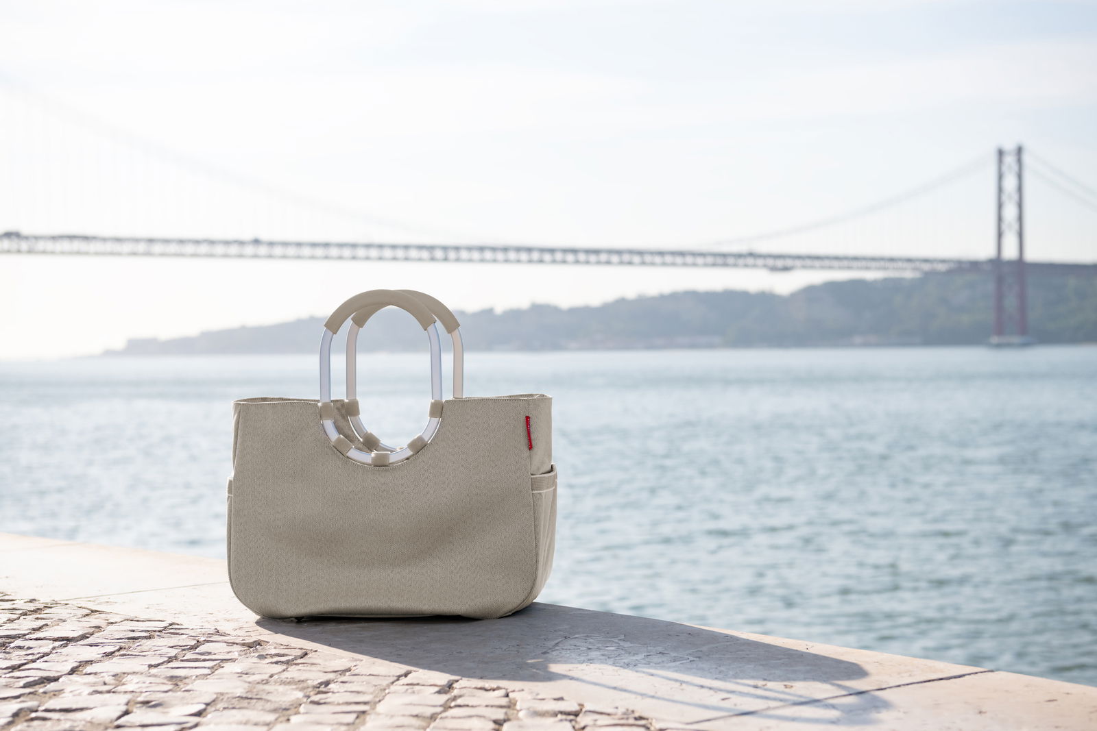 Beige Tasche mit ovalen Griffen, platziert auf einem gepflasterten Weg mit Blick auf eine Brücke und das Wasser im Hintergrund, seitliche Perspektive.