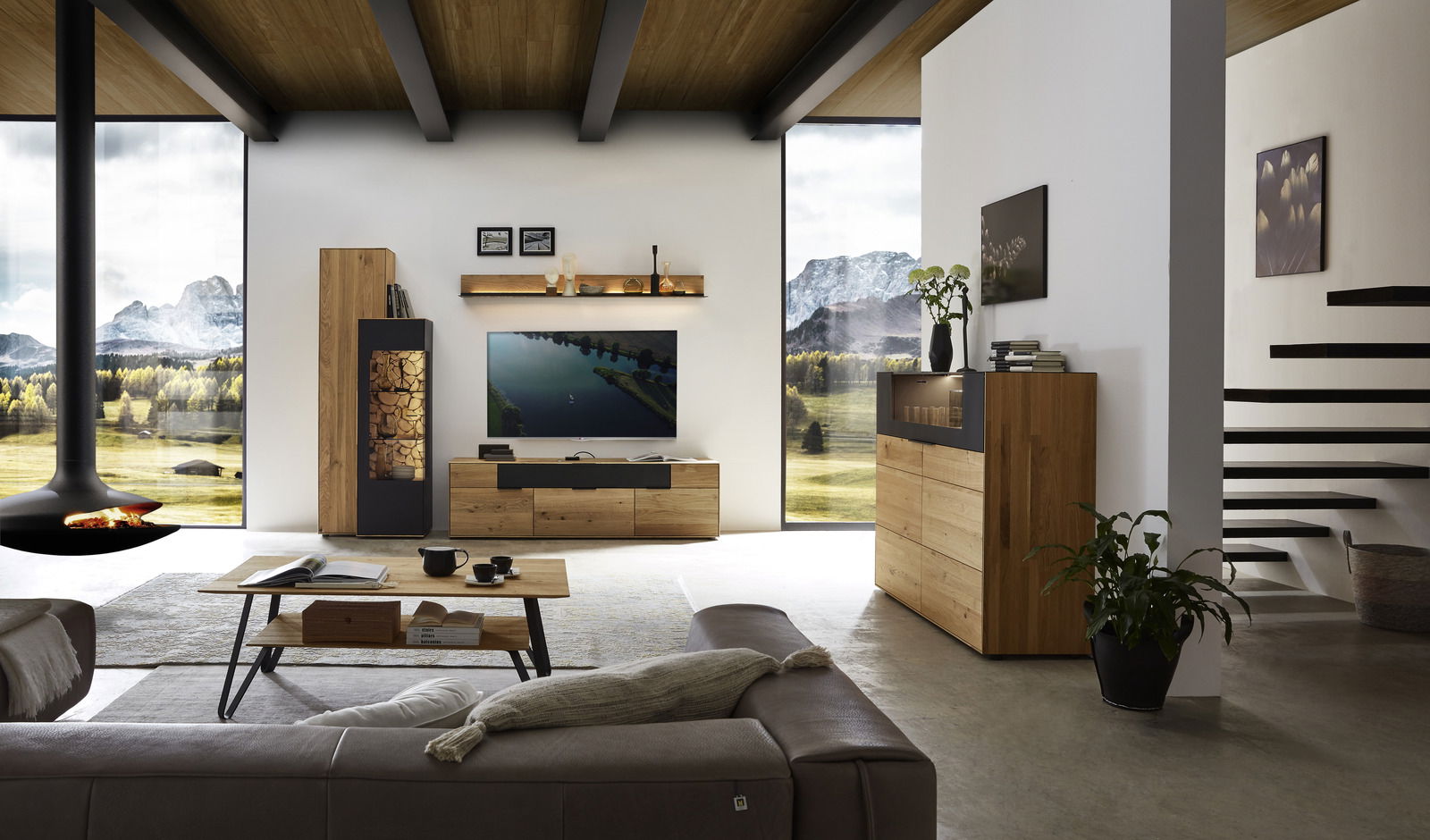 Moderne Wohnzimmeransicht mit einer Wandkombination aus Holz von Wöstmann Markenmöbel, bestehend aus einem TV-Board, einem hohen Schrank und einem Regal, mit Blick auf eine malerische Landschaft durch große Fenster.