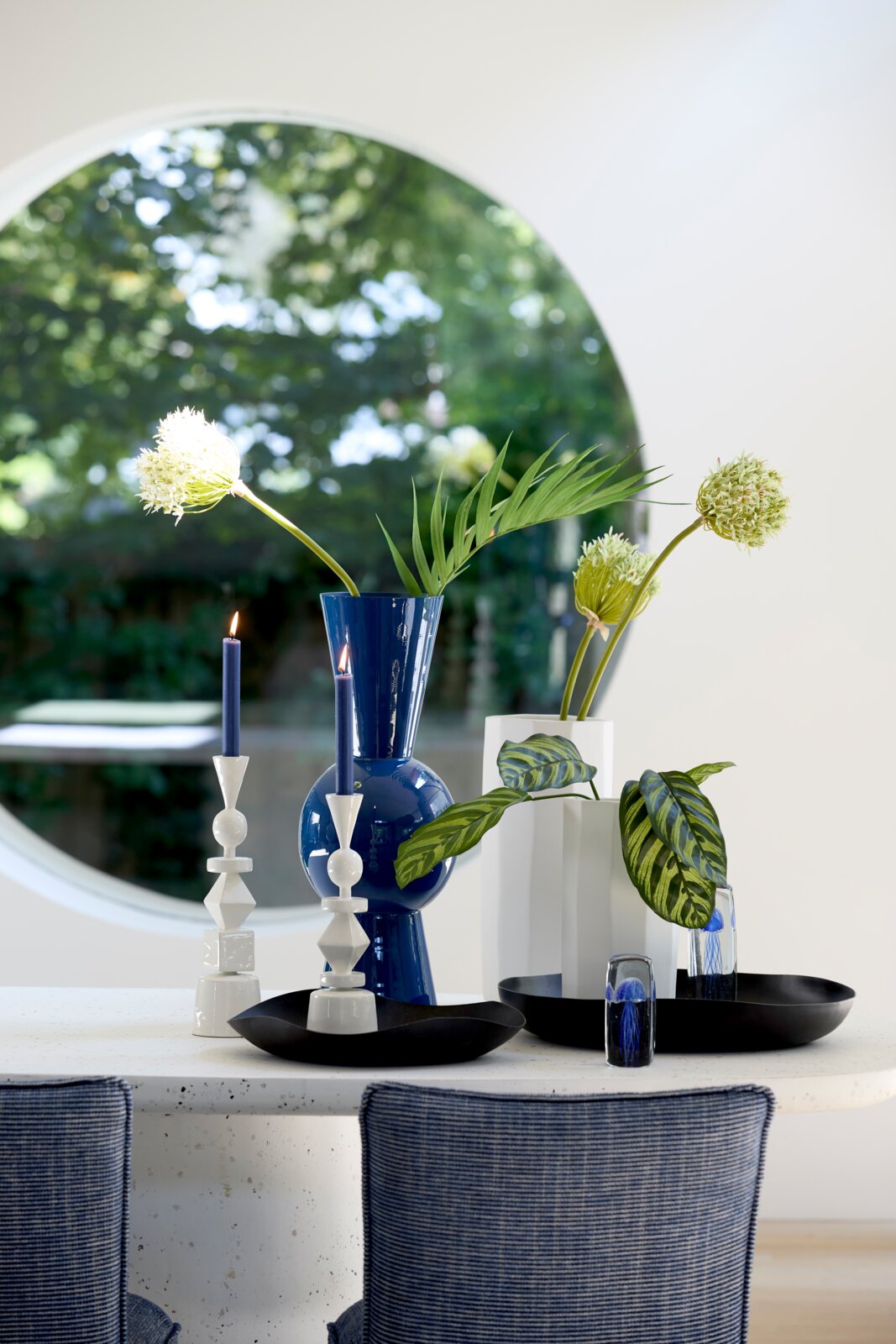 Frontaal aanzicht van een stijlvolle eettafel met kobaltblauwe vaas, witte kandelaars, zwarte schalen en bloemstukken voor een groot rond raam.