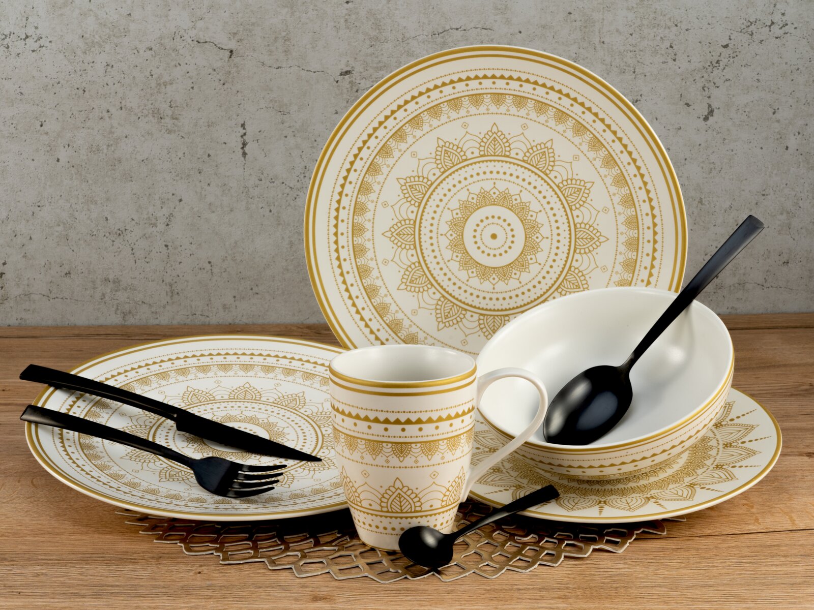Creatable Kombiservice 8-tlg MANDALA GOLD Elegantes weiß-goldenes Geschirrset mit Mandala-Ornamenten (Teller, Schale und Tasse) mit schwarzem Besteck auf Holztisch vor Betonwand, frontal leicht von oben fotografiert