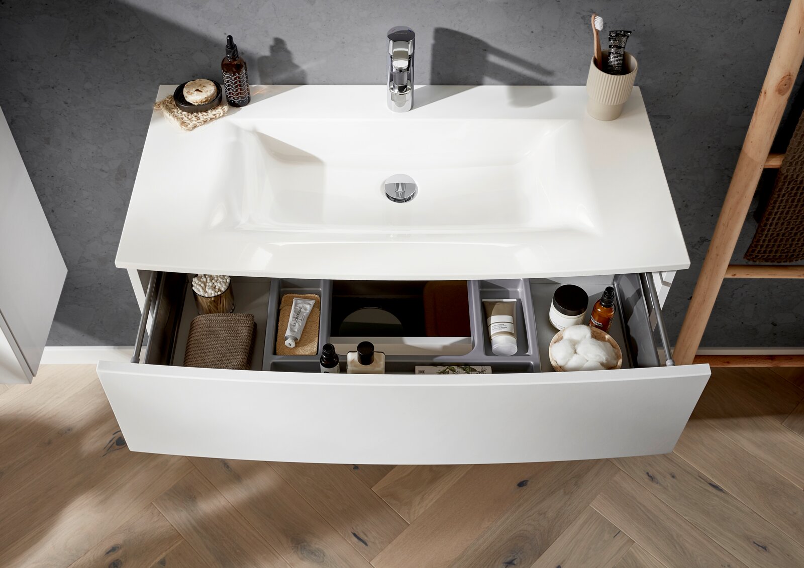 Modernes weißes Bad-Waschbecken mit integriertem Waschtisch und geöffneter Schublade mit Organizer und Pflegeartikeln, Ansicht leicht von oben/Frontperspektive.