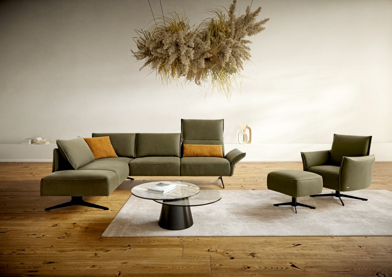 Koinor Hoekbank VINETO Moderne Polsterrundecke in Olivgrün mit passendem Sessel und Hocker, aus der Perspektive von vorne links betrachtet. Im Hintergrund eine minimalistische Dekoration und ein hängendes Pflanzenarrangement.