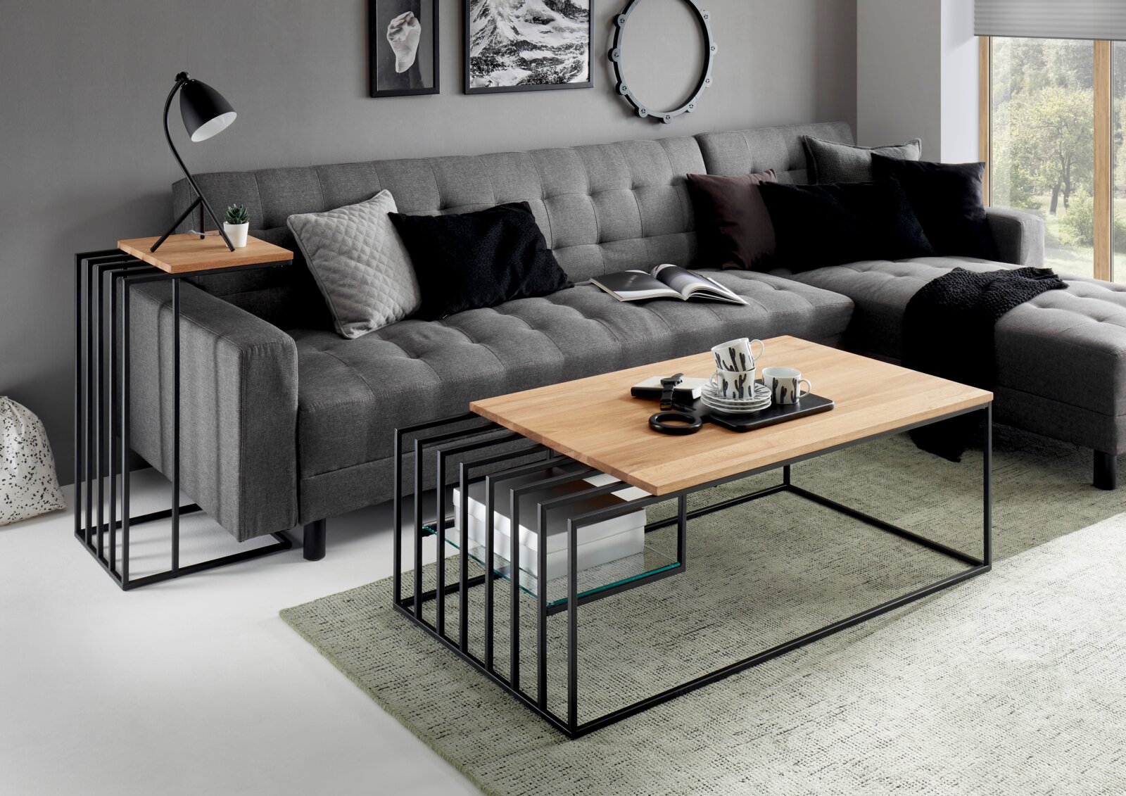 Z2 Beistelltisch JUBA Wohnzimmer-Setting mit modernem Couchtisch aus heller Holzplatte und schwarzem Metallgestell mit Ablage, davor ein graues Ecksofa; Perspektive: leicht seitliche Frontansicht aus Augenhöhe.