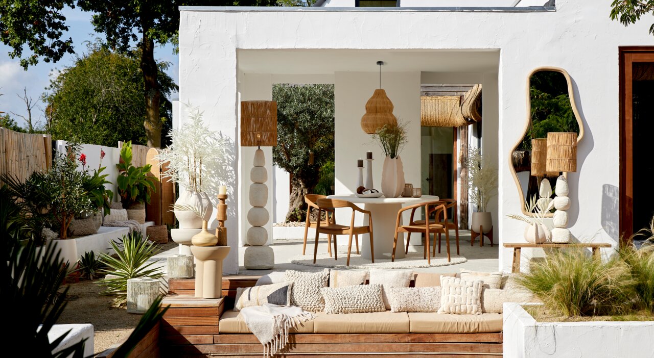 Ein stilvoller Außenbereich mit einem modernen weißen Gebäude, umgeben von üppigem Grün. Im Vordergrund befindet sich eine gemütliche Sitzgelegenheit mit mehreren dekorativen Kissen, darunter das Kissen HUMADA in Bouclé-Optik. Die Perspektive zeigt den Be