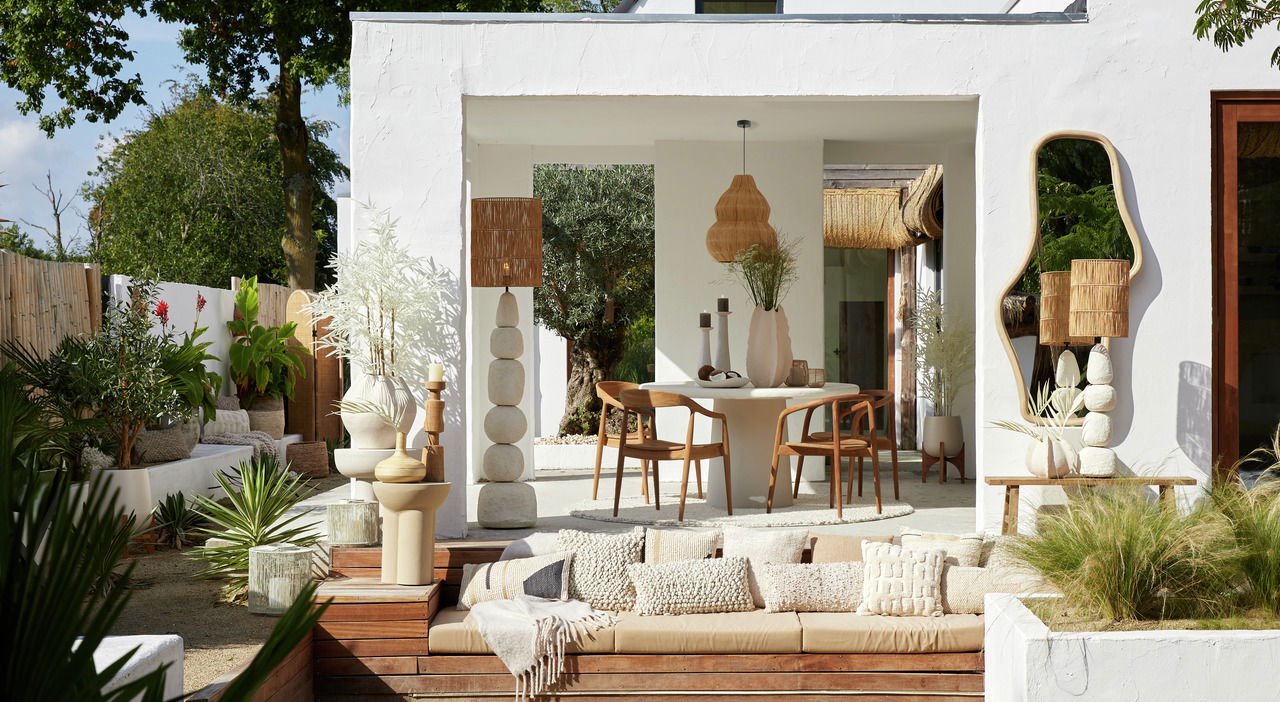 Light&Living Kissen 60x30 cm HUMADA bouclé HUMADA Ein stilvoller Außenbereich mit einem modernen weißen Gebäude, umgeben von üppigem Grün. Im Vordergrund befindet sich eine gemütliche Sitzgelegenheit mit mehreren dekorativen Kissen, darunter das Kissen HUMADA in Bouclé-Optik. Die Perspektive zeigt den Be