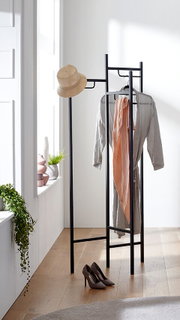 Schwarzer Metall-Kleiderständer mit Hut, Kleiderbügel mit grauem Bademantel und rosa Handtuch in hellem Raum, frontale Perspektive.