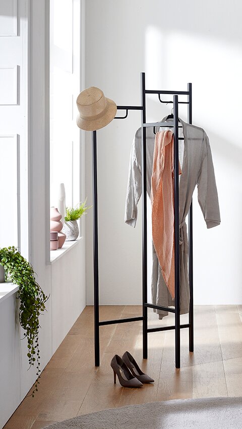 Schwarzer Metall-Kleiderständer mit Hut, Kleiderbügel mit grauem Bademantel und rosa Handtuch in hellem Raum, frontale Perspektive.