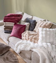 Ein gemütliches Wohnzimmer mit einer beigen Couch, auf der mehrere Felldecken und Kissen in verschiedenen Farben und Texturen gestapelt sind. Die Perspektive zeigt die Couch von schräg oben.