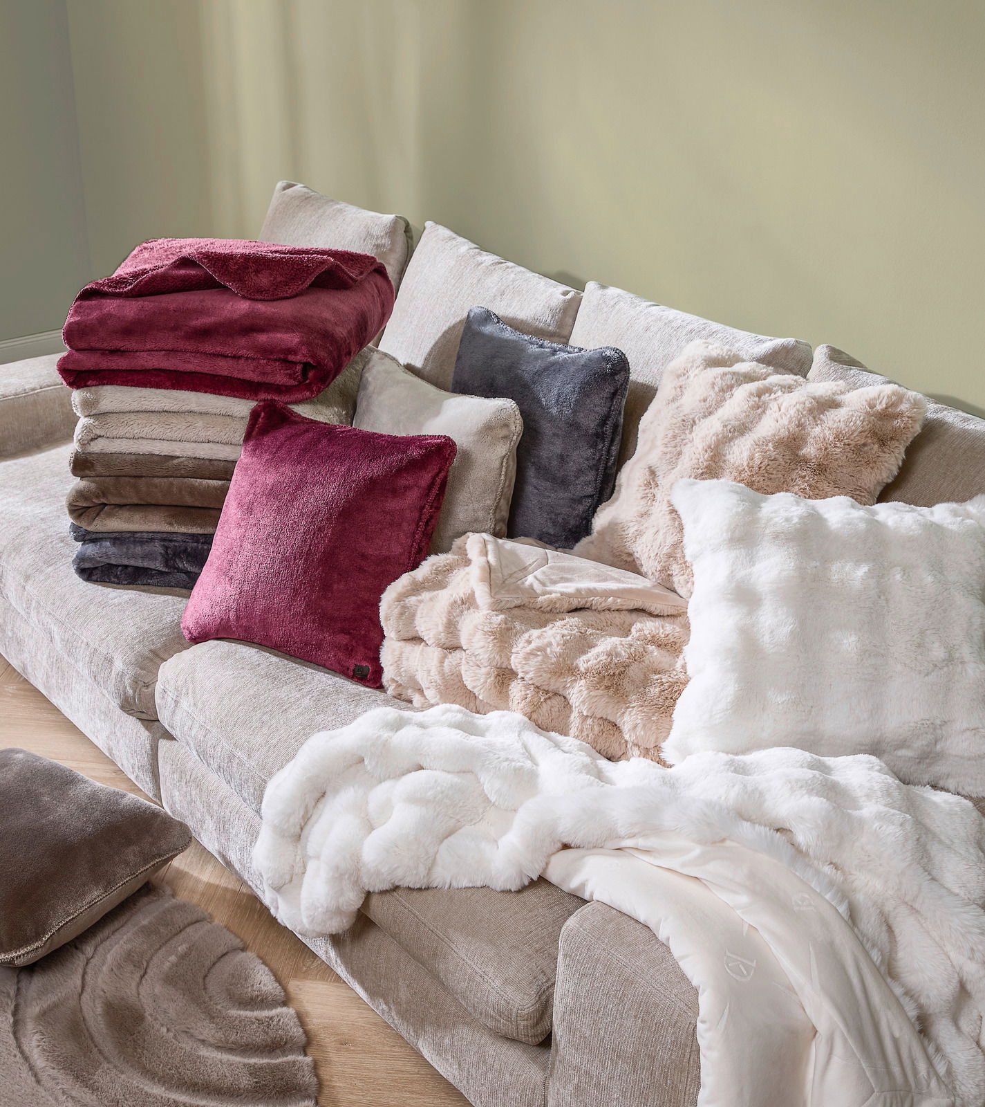 Ein gemütliches Wohnzimmer mit einer beigen Couch, auf der mehrere Felldecken und Kissen in verschiedenen Farben und Texturen gestapelt sind. Die Perspektive zeigt die Couch von schräg oben.