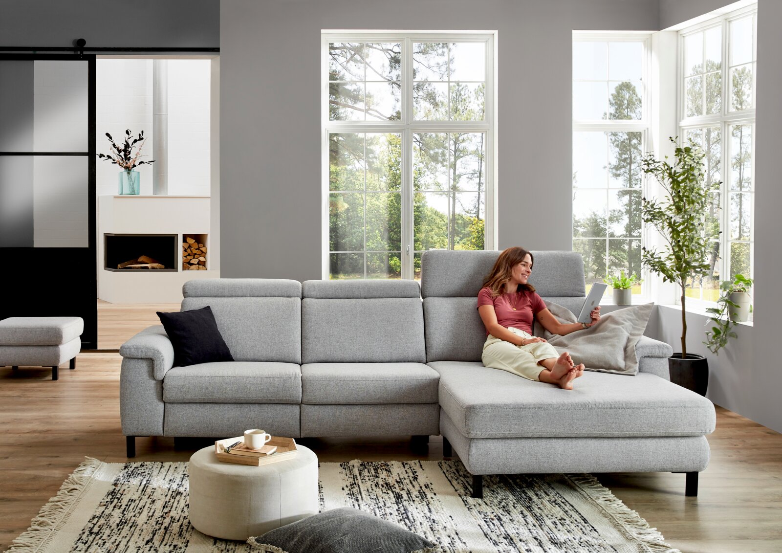 Graue Longchairkombination in einem modernen Wohnzimmer, seitliche Perspektive, mit einer Frau, die auf dem Sofa sitzt und ein Tablet benutzt.