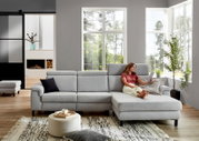 Graue Longchairkombination in einem modernen Wohnzimmer, seitliche Perspektive, mit einer Frau, die auf dem Sofa sitzt und ein Tablet benutzt.