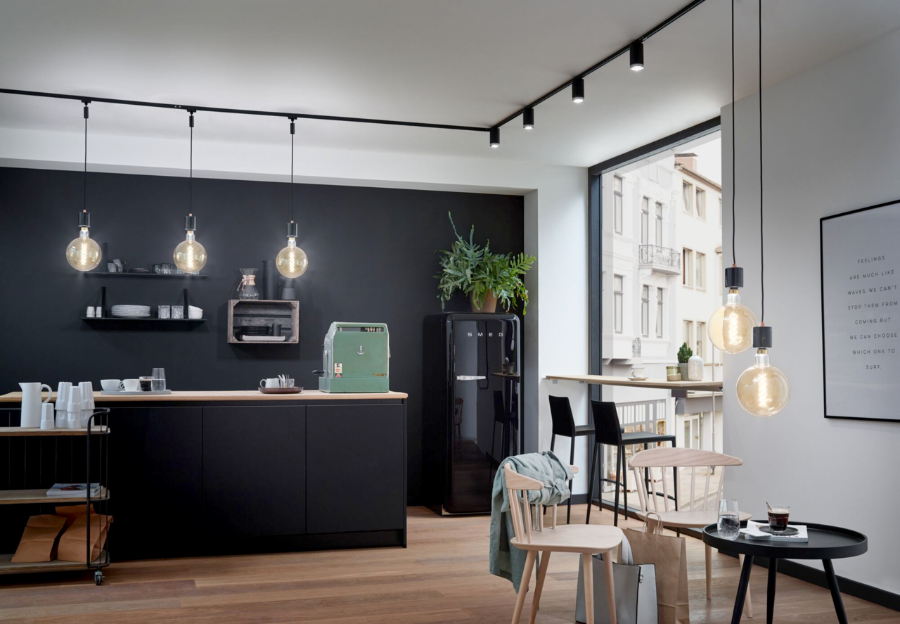 Moderne Küche mit schwarzen Hängelampen an einer Schiene, die von der Decke hängt. Perspektive von vorne auf die Küche.