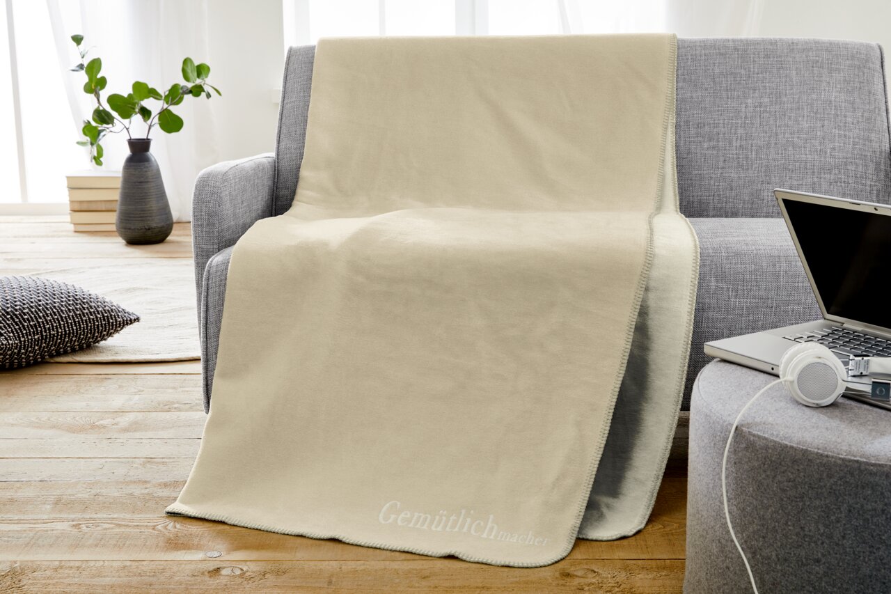 Gemütliche beige Jacquard-Decke auf einem grauen Sofa, mit einem Laptop und Kopfhörern auf einem Beistelltisch, aus der Perspektive eines Wohnzimmers.