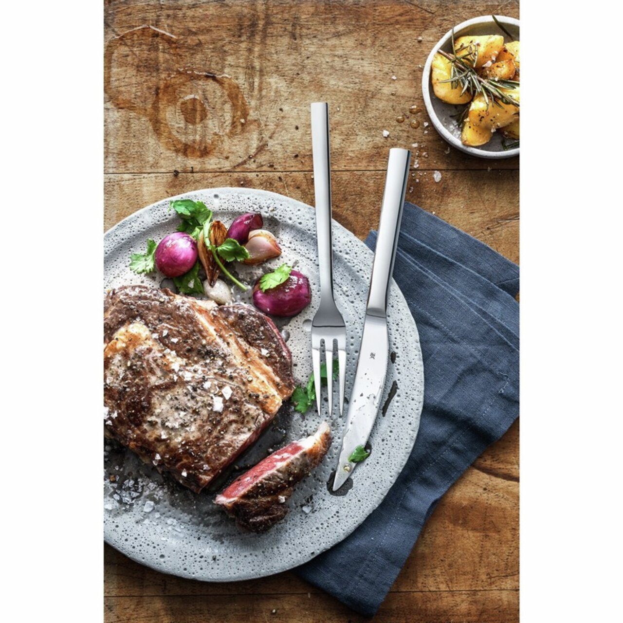 Steakbesteck Nuova auf einem rustikalen Holztisch, neben einem Teller mit saftigem Steak und Gemüse, aus der Vogelperspektive fotografiert.