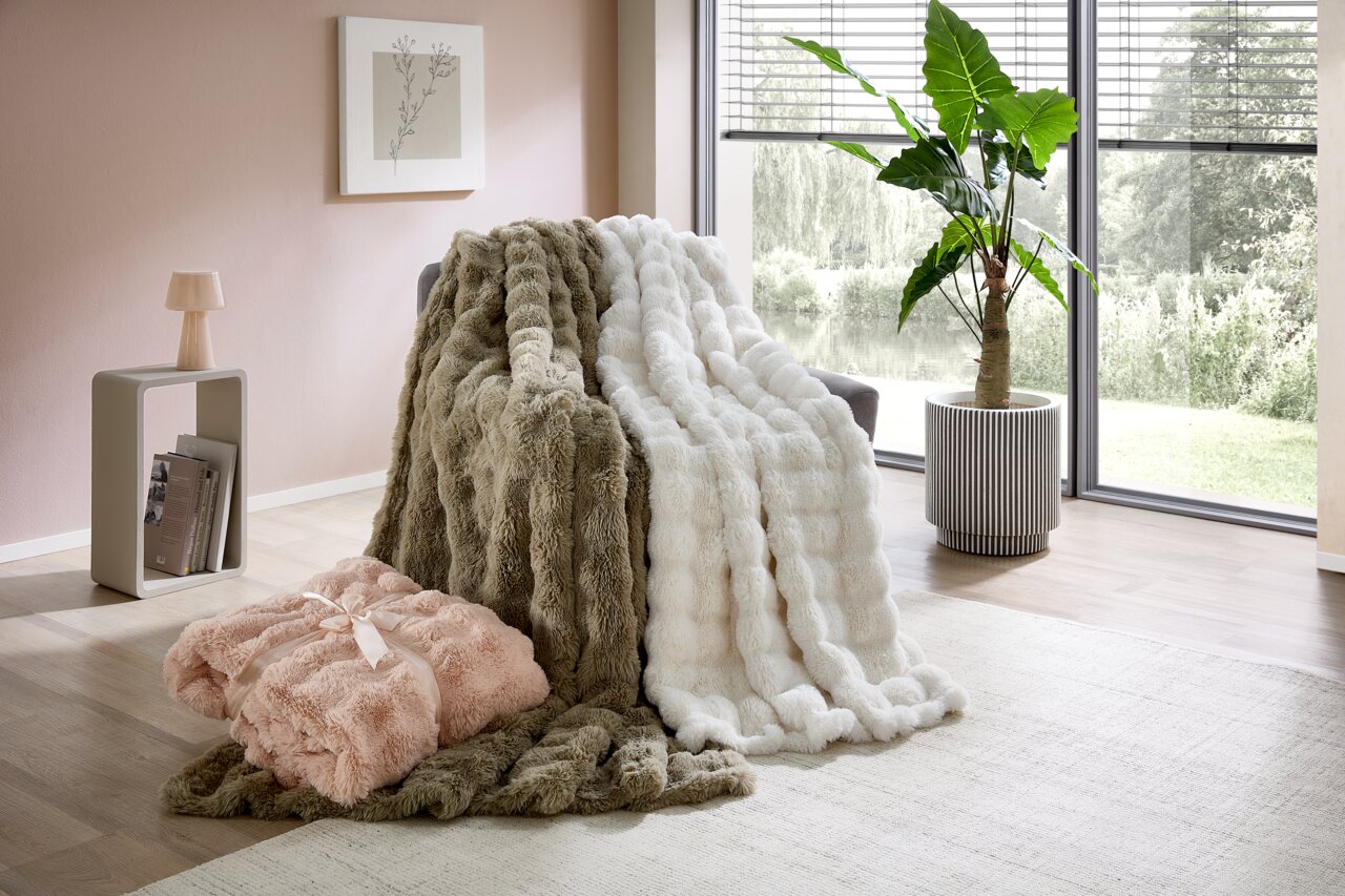 Gezellige bontdekens in verschillende kleuren, gedrapeerd over een fauteuil in een moderne woonkamer met uitzicht op de tuin.