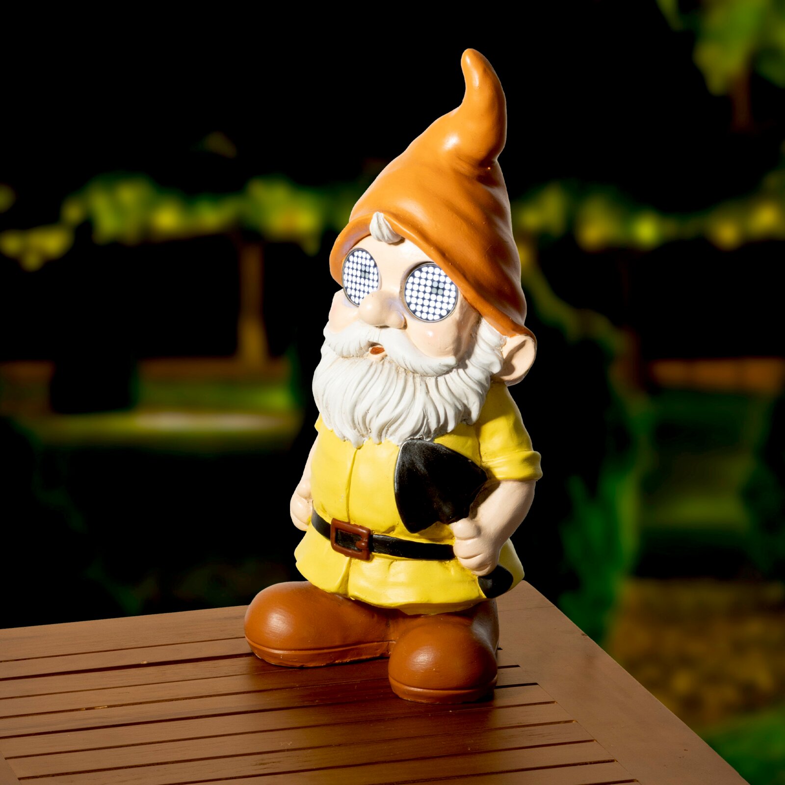 Solarbetriebener Gartenzwerg aus Polyresin mit orangefarbener Mütze und leuchtenden Augen, auf einem Holztisch stehend, bei Nacht fotografiert.