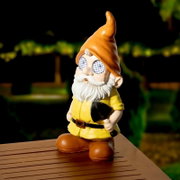Solarbetriebener Gartenzwerg aus Polyresin mit orangefarbener Mütze und leuchtenden Augen, auf einem Holztisch stehend, bei Nacht fotografiert.