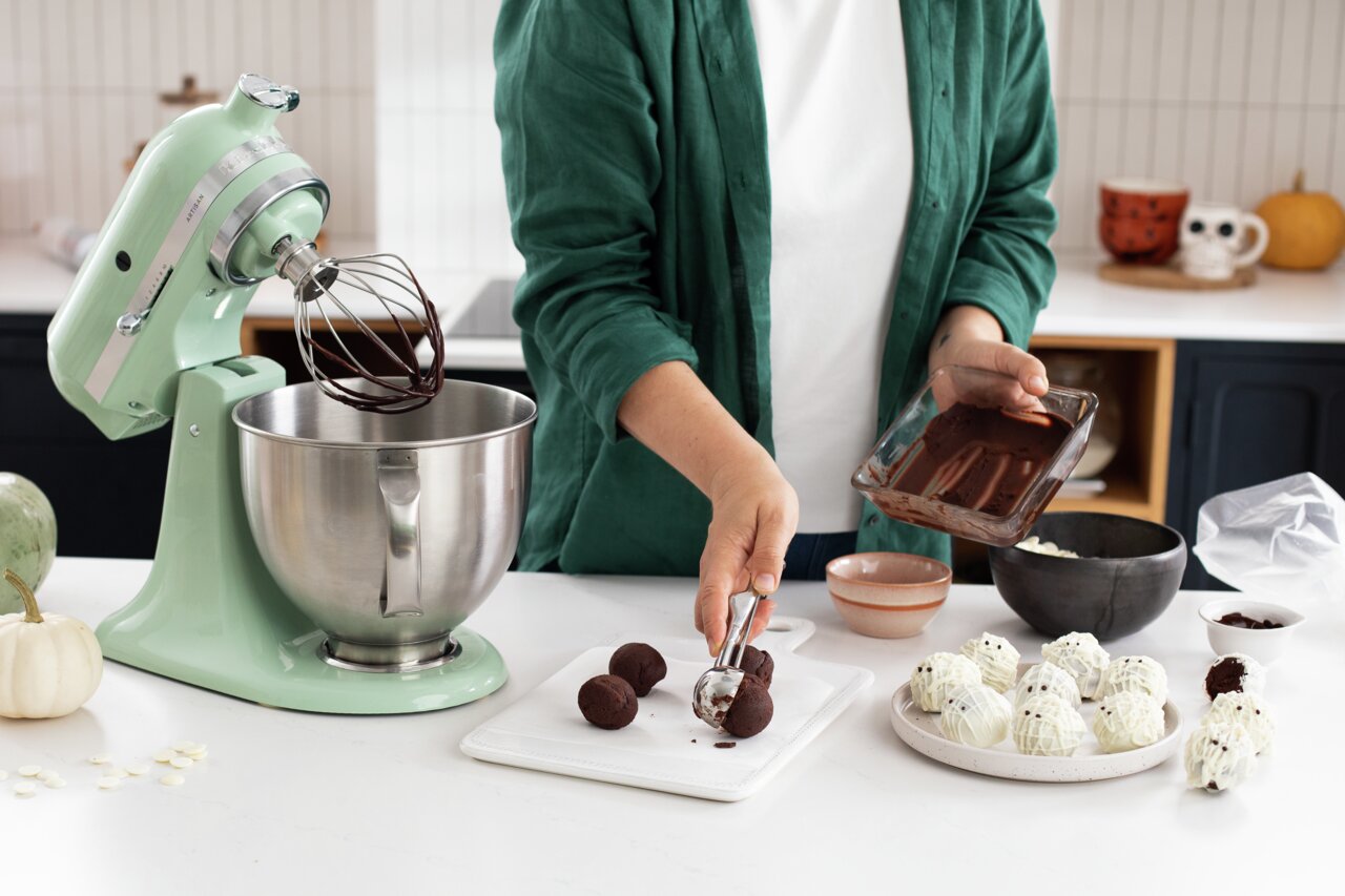 Küchenmaschine Artisan in mintgrün auf einer Arbeitsfläche, mit einer Schüssel und einem Schneebesenaufsatz, während eine Person Schokoladenkugeln formt. Perspektive von der Seite.