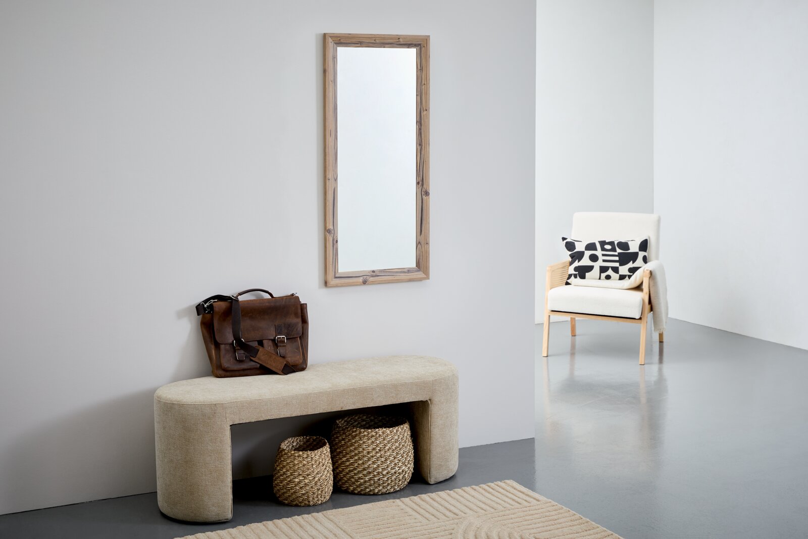 Rechteckiger Spiegel mit Holzrahmen an einer Wand, im Vordergrund eine beige Bank mit brauner Ledertasche und zwei geflochtenen Körben, seitliche Perspektive.