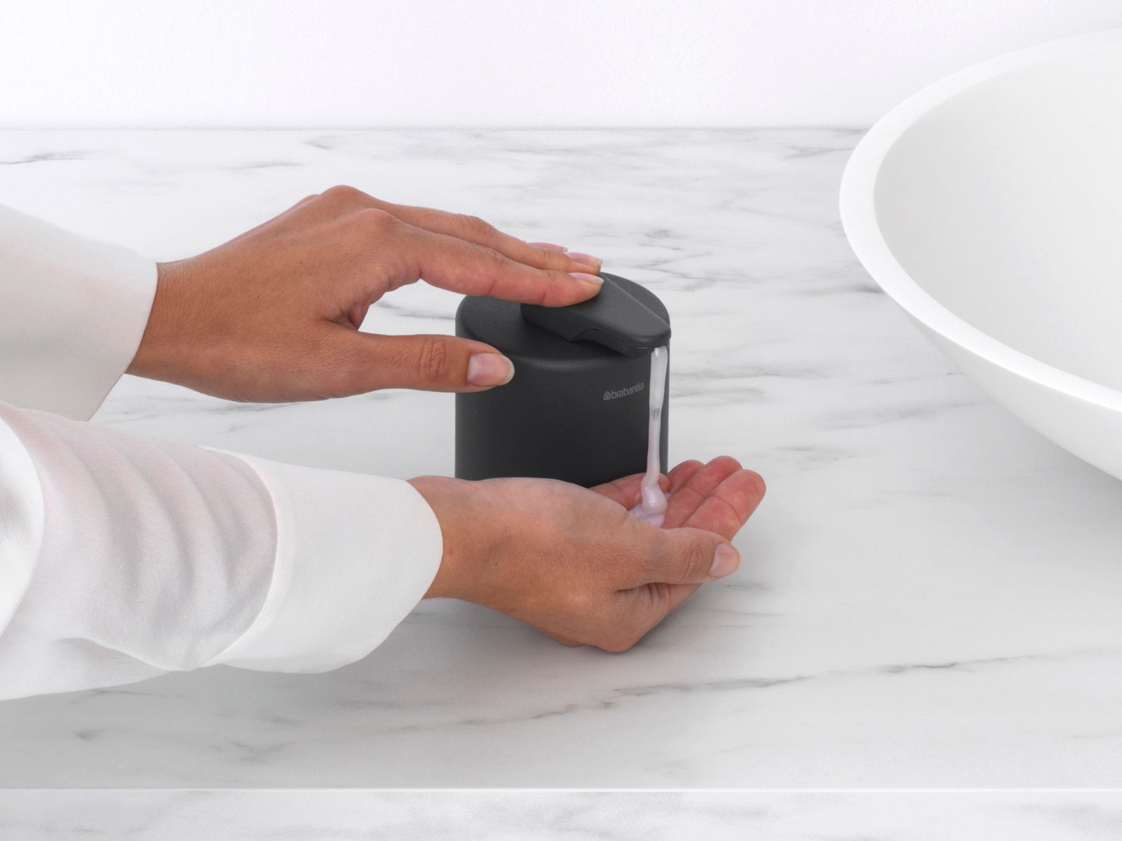 Schwarzer Seifenspender auf einer Marmoroberfläche, von der Seite gesehen, mit einer Hand, die Seife entnimmt.