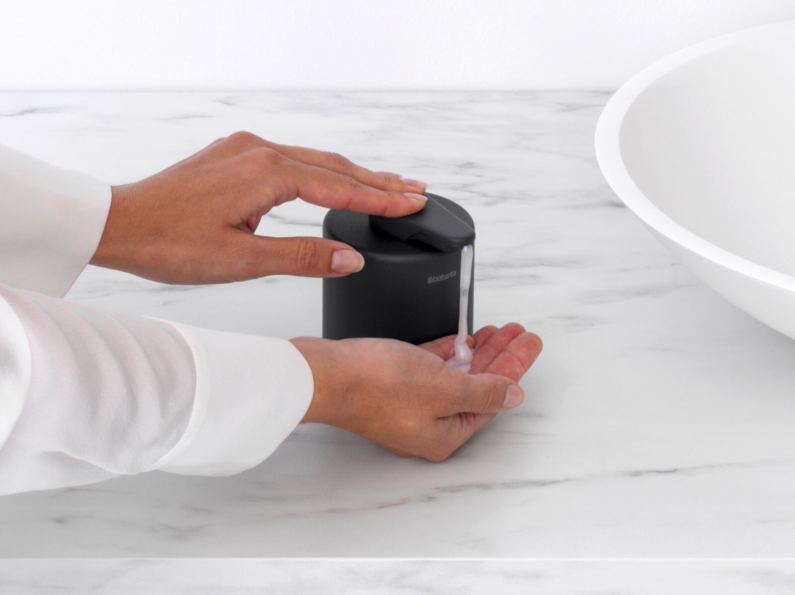Zwarte zeepdispenser op een marmeren oppervlak, gezien vanaf de zijkant, met een hand die zeep uit de dispenser haalt.
