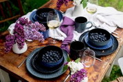 Schwarz-blau gesprenkeltes Geschirr-Set mit Schalen voller Blaubeeren, Tassen und Weingläsern auf rustikalem Holztisch mit weißen und violetten Stoffservietten sowie Fliederblüten, fotografiert aus schräger Draufsicht.
