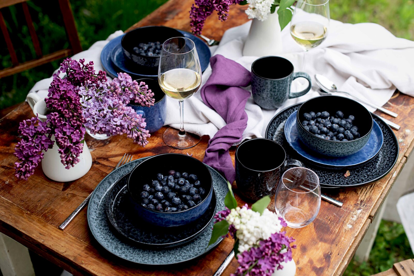 Schwarz-blau gesprenkeltes Geschirr-Set mit Schalen voller Blaubeeren, Tassen und Weingläsern auf rustikalem Holztisch mit weißen und violetten Stoffservietten sowie Fliederblüten, fotografiert aus schräger Draufsicht.