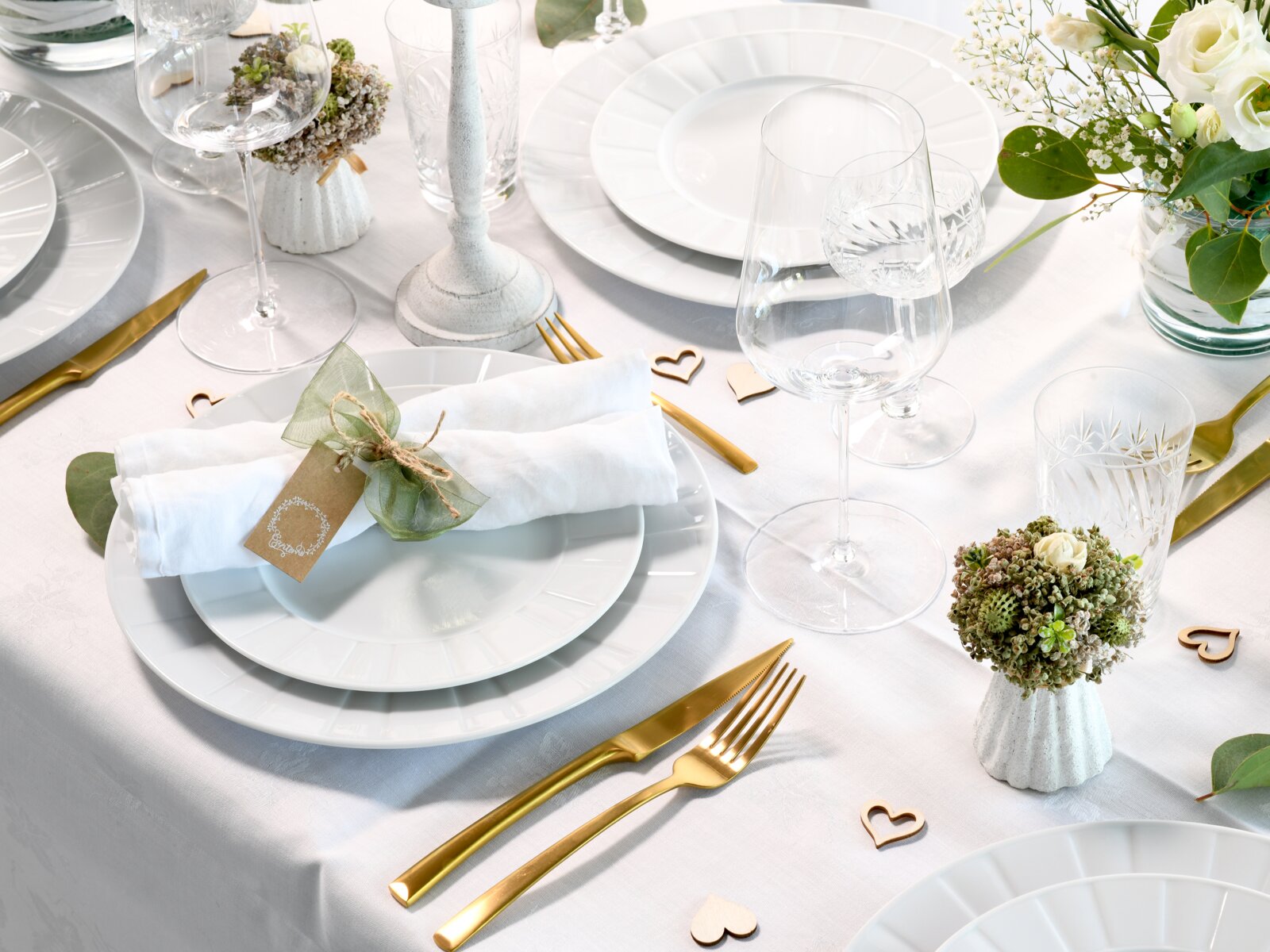 Festlich gedeckter Tisch in heller Draufsicht/Schrägansicht: weiße Teller mit gerollter Serviette und Namensschild, goldfarbenes Besteck, Kristallgläser, weiße Kerzenhalter, kleine Blumenarrangements und Herz-Deko auf weißer Tischdecke.