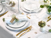 Festlich gedeckter Tisch in heller Draufsicht/Schrägansicht: weiße Teller mit gerollter Serviette und Namensschild, goldfarbenes Besteck, Kristallgläser, weiße Kerzenhalter, kleine Blumenarrangements und Herz-Deko auf weißer Tischdecke.