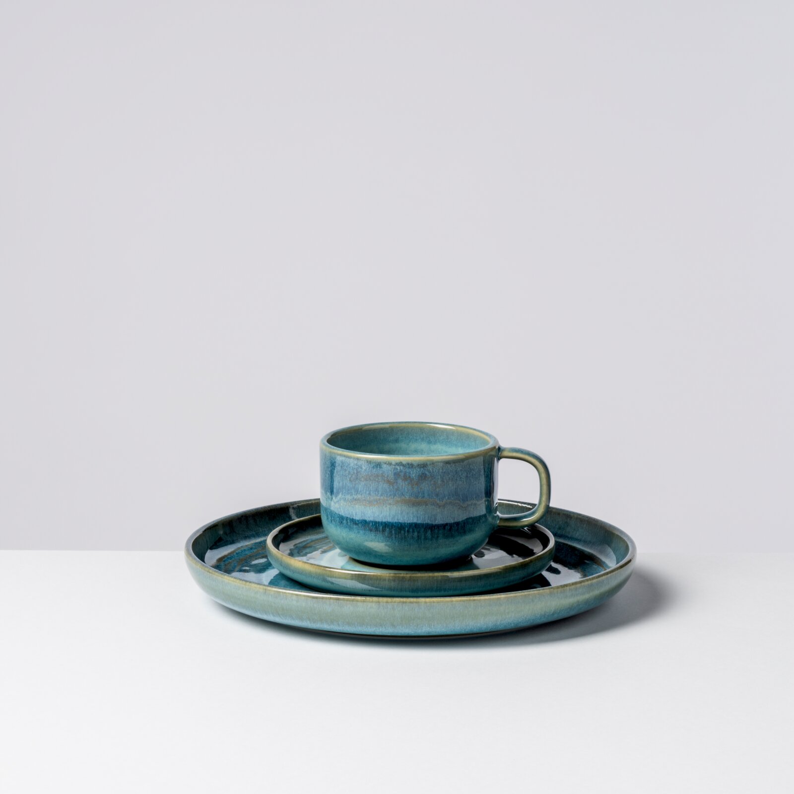 Blau glasierte Keramik-Tasse mit Henkel auf passender Untertasse und großem Teller, Studioaufnahme vor hellem Hintergrund, Frontansicht leicht von rechts