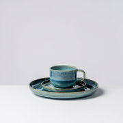 Blau glasierte Keramik-Tasse mit Henkel auf passender Untertasse und großem Teller, Studioaufnahme vor hellem Hintergrund, Frontansicht leicht von rechts