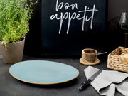 Hellblauer, runder Keramikteller mit braunem Rand auf dunkler Tischplatte, im Hintergrund schwarze Tafel mit Schriftzug „bon appetit“, daneben Kräutertopf, Brotkorb und Messer – Perspektive: leicht von vorne/seitlich aus Tischhöhe.
