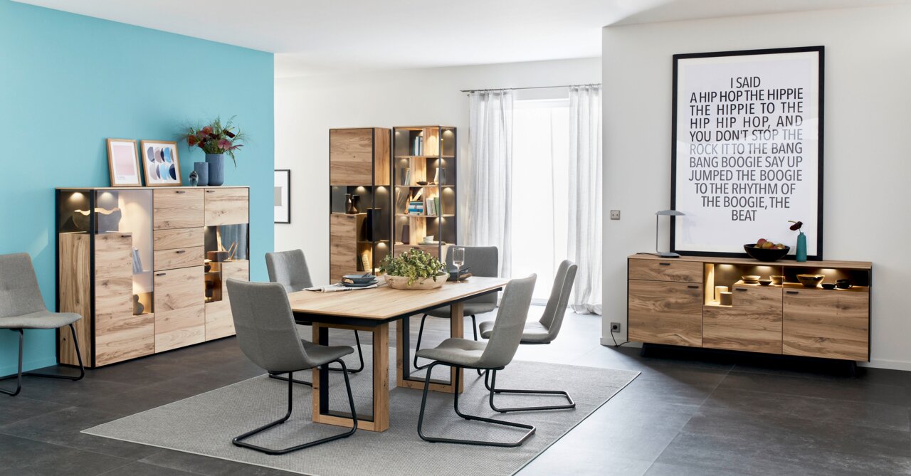 Modernes Highboard aus Holz mit Glasvitrinen, dekoriert mit Pflanzen und Kunstwerken, in einem stilvollen Esszimmer. Perspektive: Frontalansicht.
