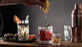 Ein Set aus vier Schnapsgläsern auf einem Tisch, mit einem Cocktail im Vordergrund und einer Hand, die eine Zitronenscheibe darüber auspresst. Perspektive von vorne.