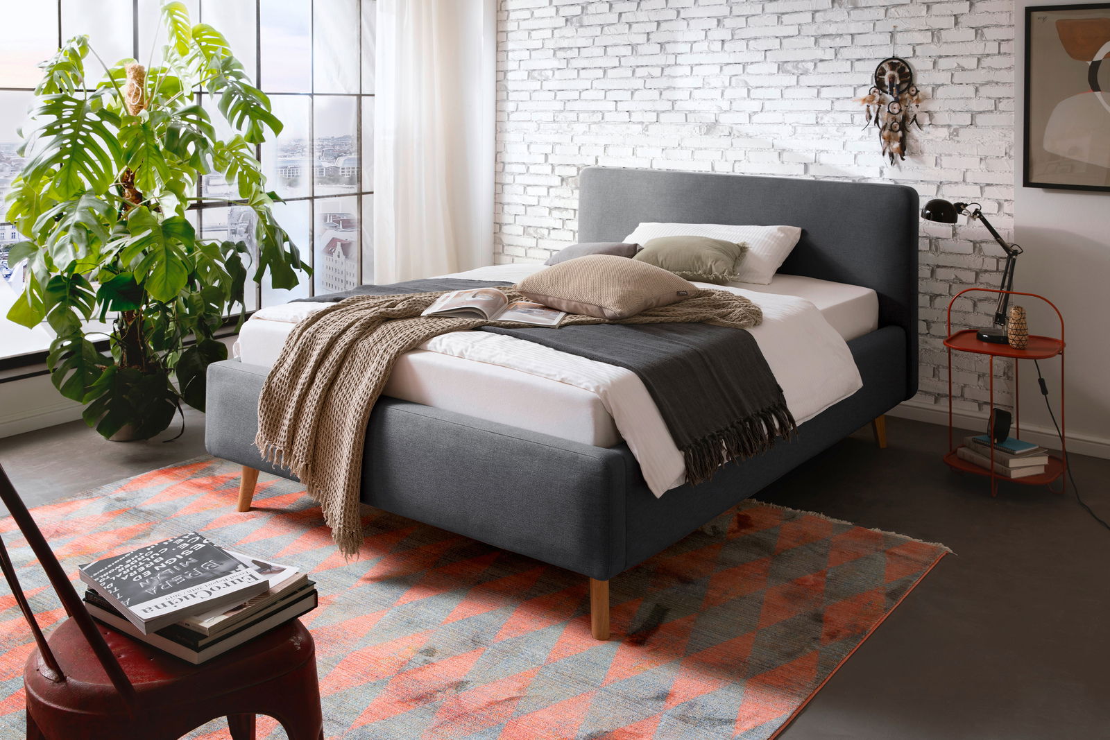 Z2 Polsterbett MATTIS Modernes Polsterbett in Grau mit Holzfüßen, dekoriert mit Kissen und Decken, in einem stilvollen Schlafzimmer mit Backsteinwand und großem Fenster, seitliche Perspektive.