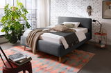 Modernes Polsterbett in Grau mit Holzfüßen, dekoriert mit Kissen und Decken, in einem stilvollen Schlafzimmer mit Backsteinwand und großem Fenster, seitliche Perspektive.