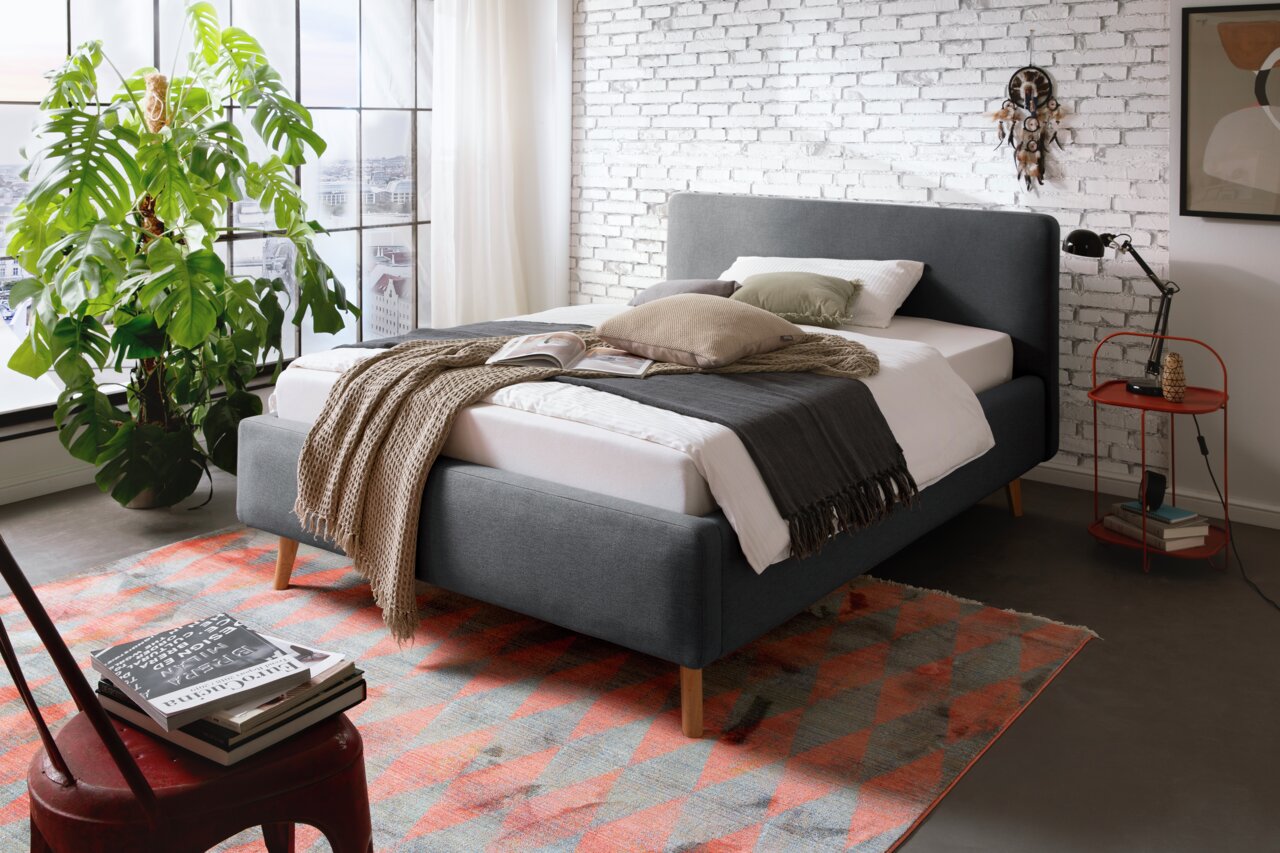 Modernes Polsterbett in Grau mit Holzfüßen, dekoriert mit Kissen und Decken, in einem stilvollen Schlafzimmer mit Backsteinwand und großem Fenster, seitliche Perspektive.