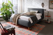 Modernes Polsterbett in Grau mit Holzfüßen, dekoriert mit Kissen und Decken, in einem stilvollen Schlafzimmer mit Backsteinwand und großem Fenster, seitliche Perspektive.