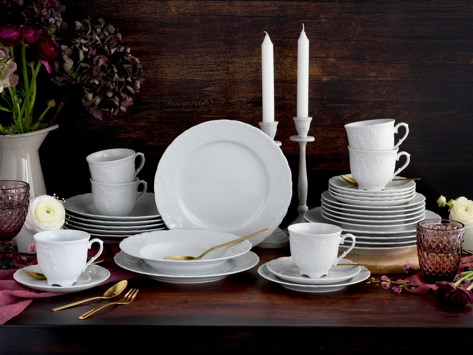 Frontale Tischansicht eines weißen Porzellan-Geschirrsets mit geprägtem Relief (Teller, Schalen, Tassen mit Untertassen), arrangiert auf dunklem Holztisch mit goldfarbenem Besteck, Kerzenständern, Blumen und rosafarbenen Gläsern vor rustikalem Holz-Hinter