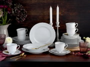 Frontale Tischansicht eines weißen Porzellan-Geschirrsets mit geprägtem Relief (Teller, Schalen, Tassen mit Untertassen), arrangiert auf dunklem Holztisch mit goldfarbenem Besteck, Kerzenständern, Blumen und rosafarbenen Gläsern vor rustikalem Holz-Hinter
