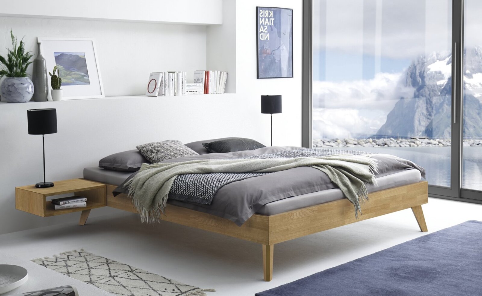Modern houten bed met zwevend effect en schuine houten poten, bekleed in grijs en versierd met een deken en kussens, in een lichte slaapkamer met grote ramen perspectief: zijaanzicht, licht van voren gezien.