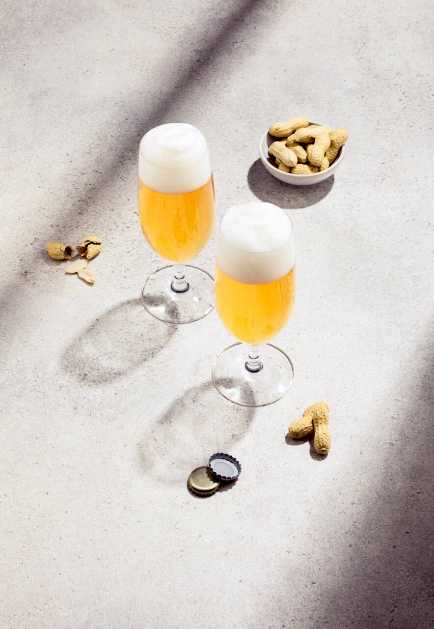 Twee biertulpen met een schuimkraag, gevuld met goudkleurig bier, op een lichte ondergrond. Op de achtergrond een schaal met pinda’s en verspreide pindaschillen. Aanzicht van bovenaf.
