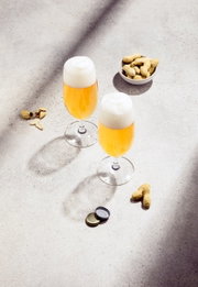Twee biertulpen met een schuimkraag, gevuld met goudkleurig bier, op een lichte ondergrond. Op de achtergrond een schaal met pinda’s en verspreide pindaschillen. Aanzicht van bovenaf.