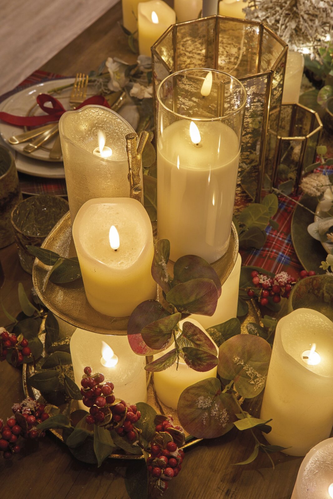 Sfeervolle collectie crèmekleurige LED-kaarsen tussen eucalyptus, rode bessen en goudkleurige windlichten, schuin van boven gefotografeerd op een feestelijk gedekte tafel.