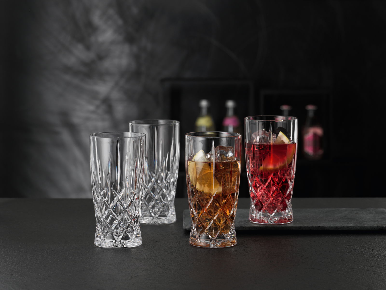 Vier elegante Softdrinkgläser aus Kristallglas im Noblesse-Design, zwei gefüllt mit Getränken und Eiswürfeln, auf einer dunklen Oberfläche vor einem unscharfen Hintergrund, seitliche Perspektive.