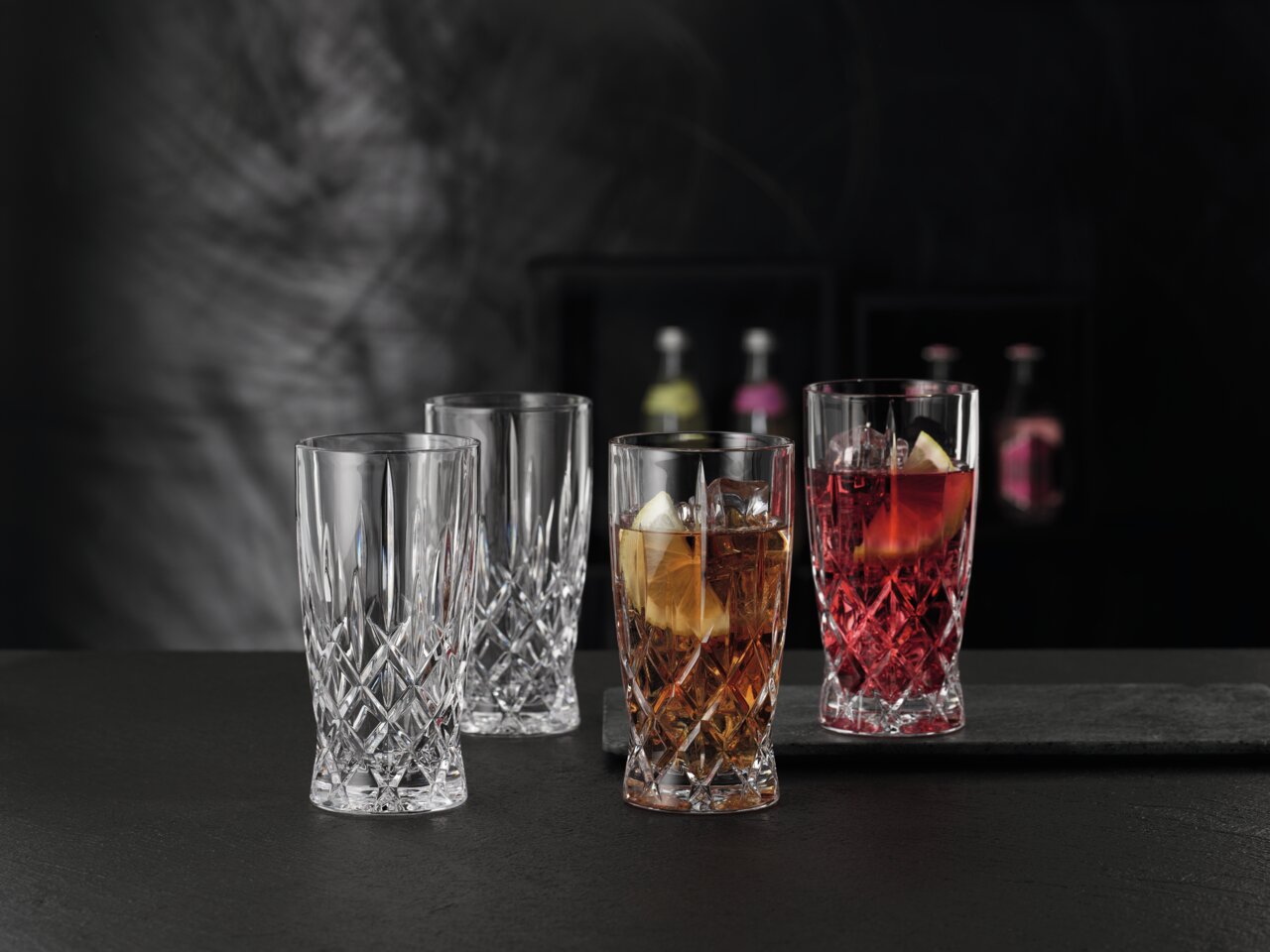 Vier elegante Softdrinkgläser aus Kristallglas im Noblesse-Design, zwei gefüllt mit Getränken und Eiswürfeln, auf einer dunklen Oberfläche vor einem unscharfen Hintergrund, seitliche Perspektive.