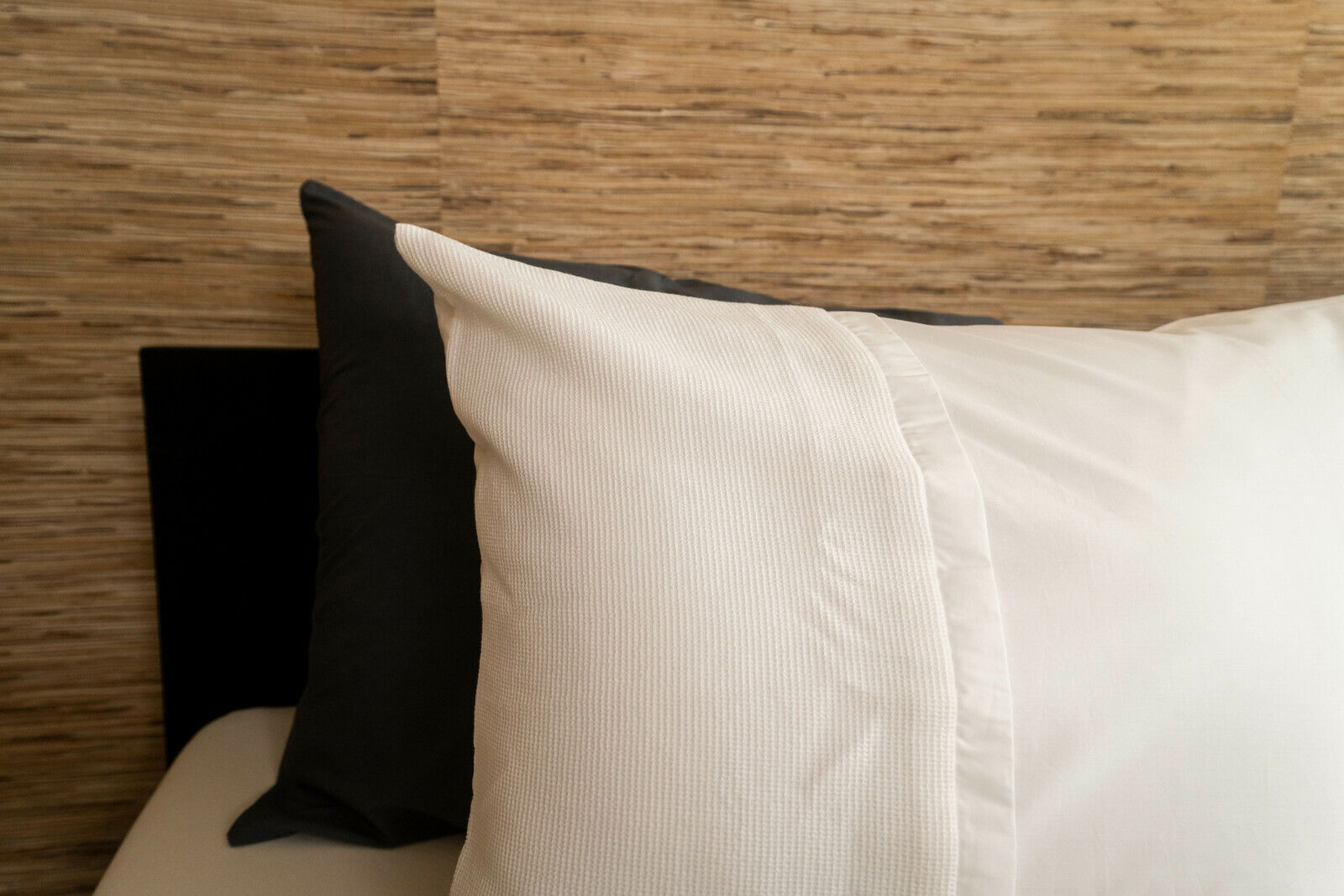 Nahaufnahme von Bettwäsche in Weiß mit strukturiertem Muster, auf einem Bett mit Holzwand im Hintergrund.