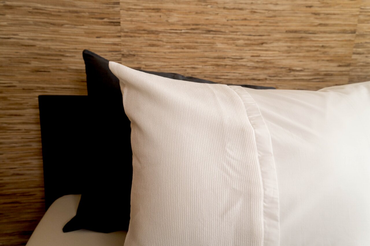 Nahaufnahme von Bettwäsche in Weiß mit strukturiertem Muster, auf einem Bett mit Holzwand im Hintergrund.